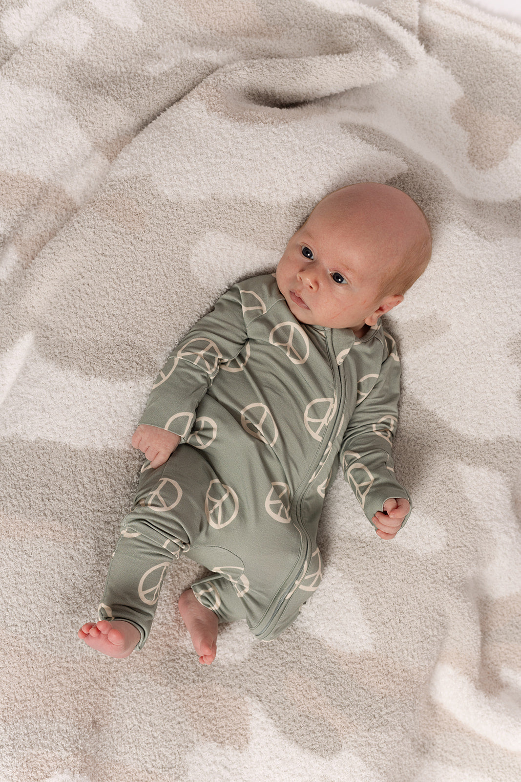 A baby lies on a soft blanket, wearing forever french baby’s Bamboo Zip Pajamas | Peace—a sage green, breathable onesie with a white peace sign pattern. The baby gazes up calmly, arms relaxed at their sides.