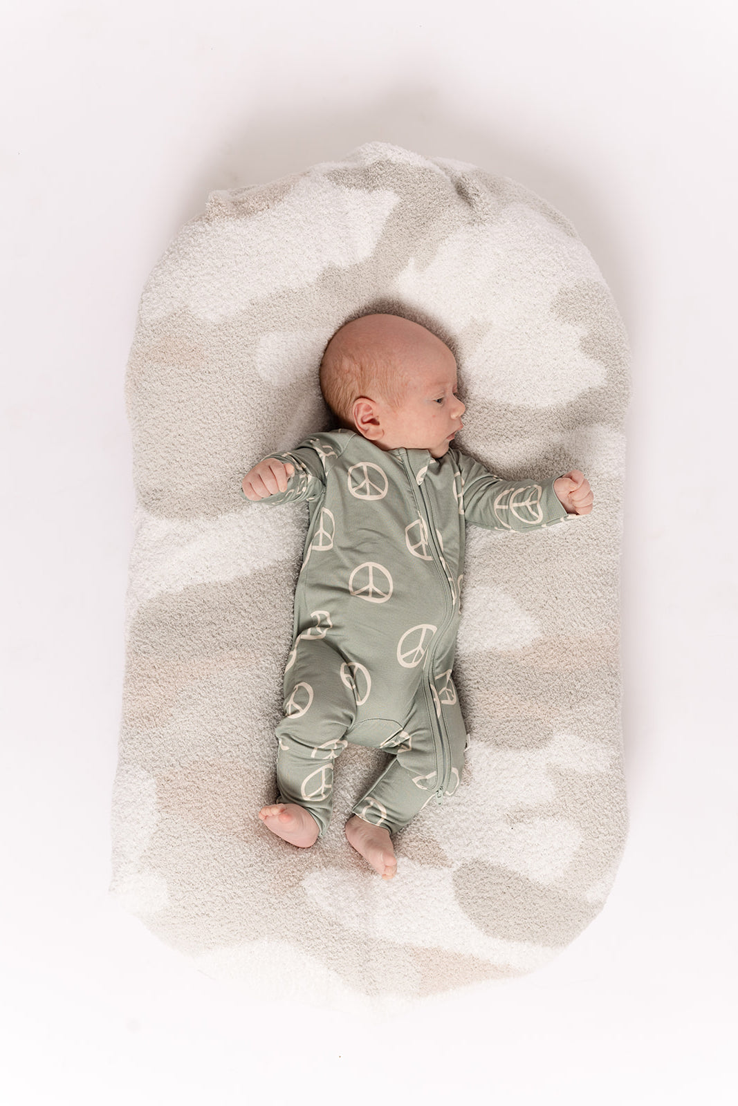 A baby wearing forever french baby's Bamboo Zip Pajamas | Peace—a green onesie with white peace signs—rests on a soft, oval, light-colored cushion against a white background.