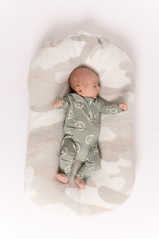 A baby wearing forever french baby's Bamboo Zip Pajamas | Peace—a green onesie with white peace signs—rests on a soft, oval, light-colored cushion against a white background.