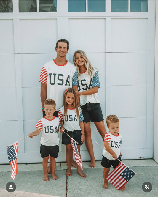 A family of five, all in forever french baby Adult Bamboo T-Shirt | Stars & Stripes and holding American flags, stands barefoot in front of a white garage door, smiling for the photo.