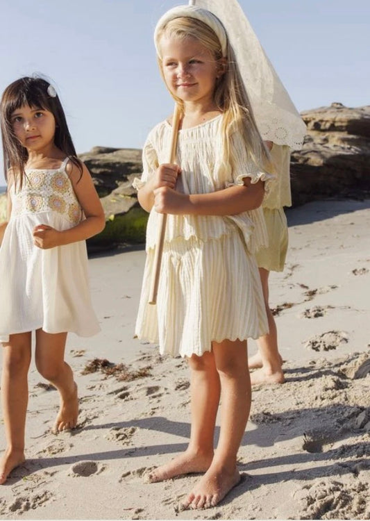 Two young girls in light dresses stand barefoot on a sandy beach. One is wearing the Rylee + Cru Smocked V Mini Skirt in Citron Stripe, crafted from 100% cotton. She holds a net as both look toward the camera, with rocks and a blue sky visible in the background.