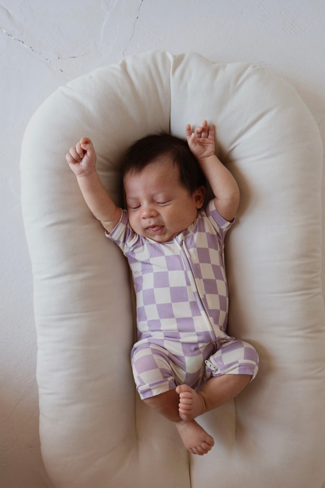 A baby wearing the forever french baby Shortie Bamboo Zip | Grape Soda sleeps peacefully on a soft, white cushion with arms raised, dressed in a lavender and white checkered hypoallergenic outfit.