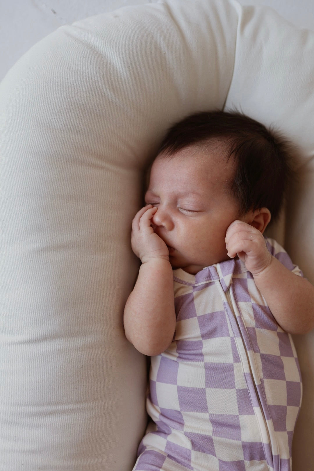 A newborn with dark hair sleeps peacefully in a cushioned, white baby lounger, wearing the forever french baby Shortie Bamboo Zip in Grape Soda—a purple and white checkered, hypoallergenic onesie—and sucking their thumb.