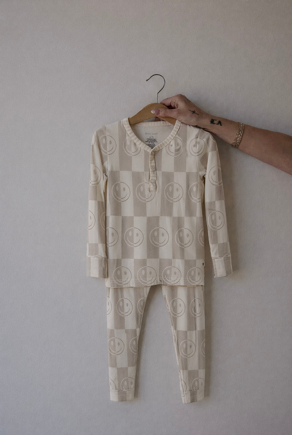 A hand holds forever french baby's Bamboo Two Piece Pajamas | Smile Checkerboard, a beige long-sleeve toddler set with a checkered pattern and smiley faces, against a plain light background.