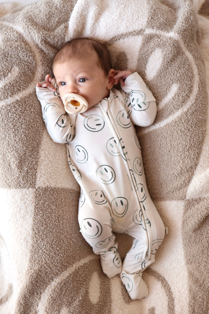 A baby in forever french baby's Bamboo Zip Pajama | Green Smiles, a breathable onesie with smiley faces, relaxes on a tan and cream patterned blanket, gazing upward with a pacifier.