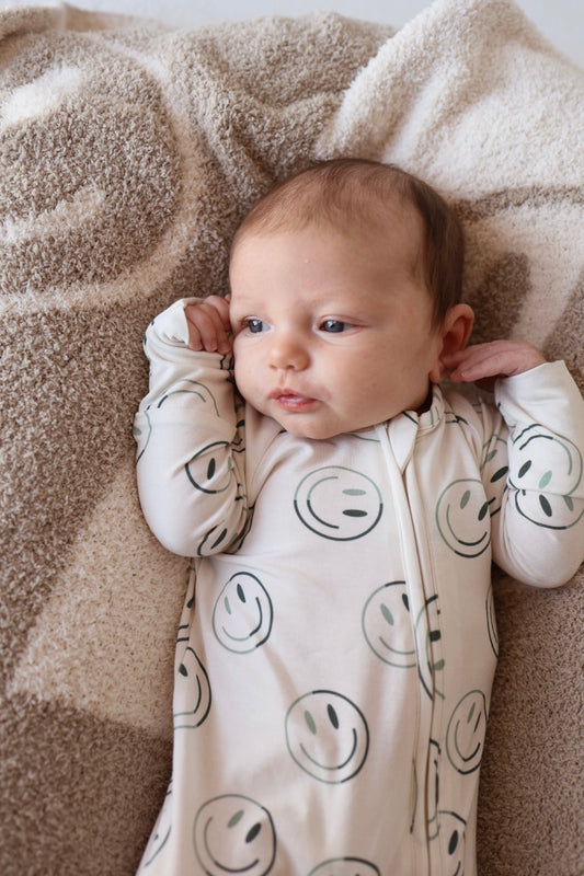 A baby wearing forever french baby's Bamboo Zip Pajama in Green Smiles lies on a soft beige and white blanket, looking off to the side with one arm bent near the head.