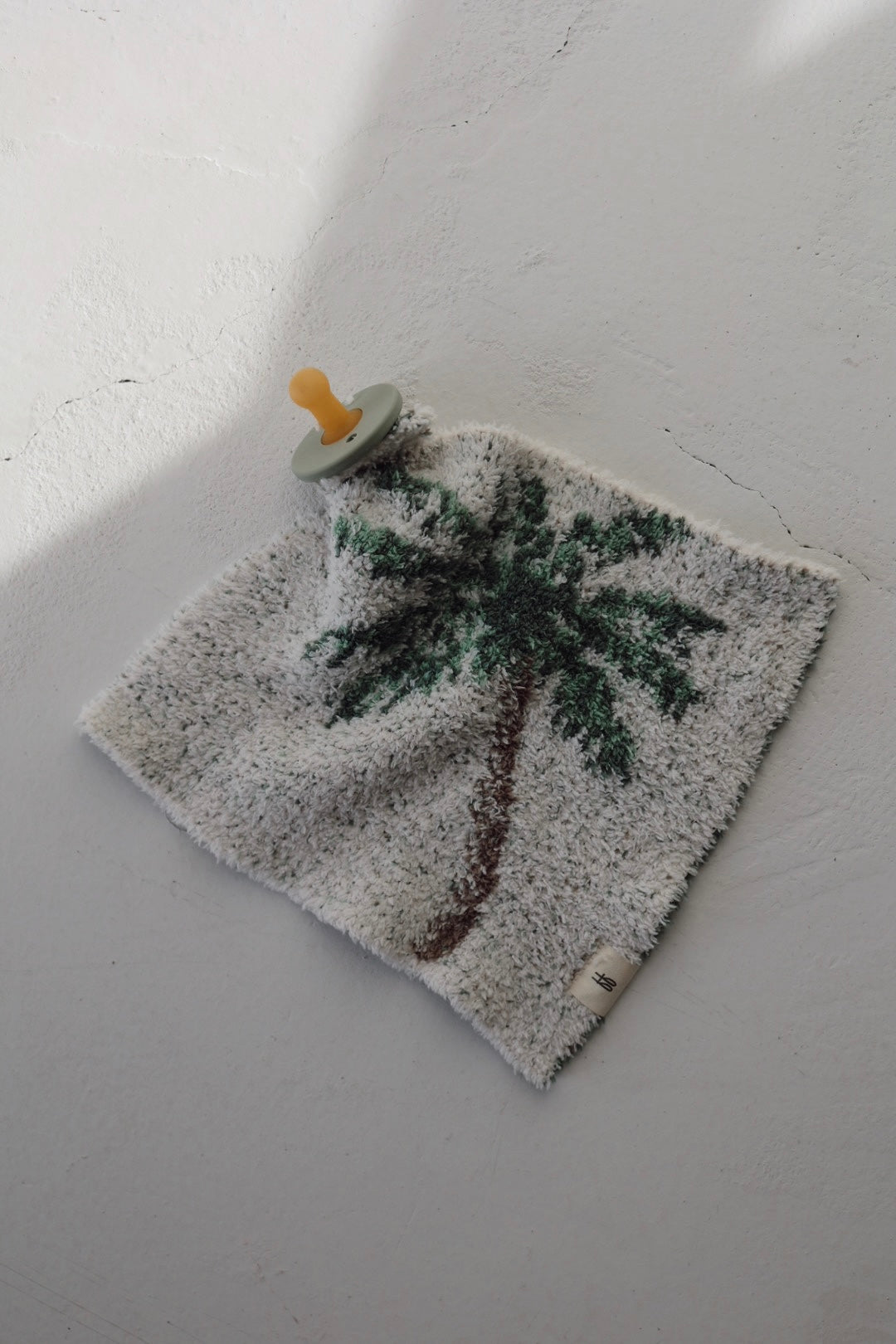The forever french baby Lovey | Palm Tree features a yellow pacifier attached to a soft, square security blanket with a palm tree design, shown on a light textured surface.