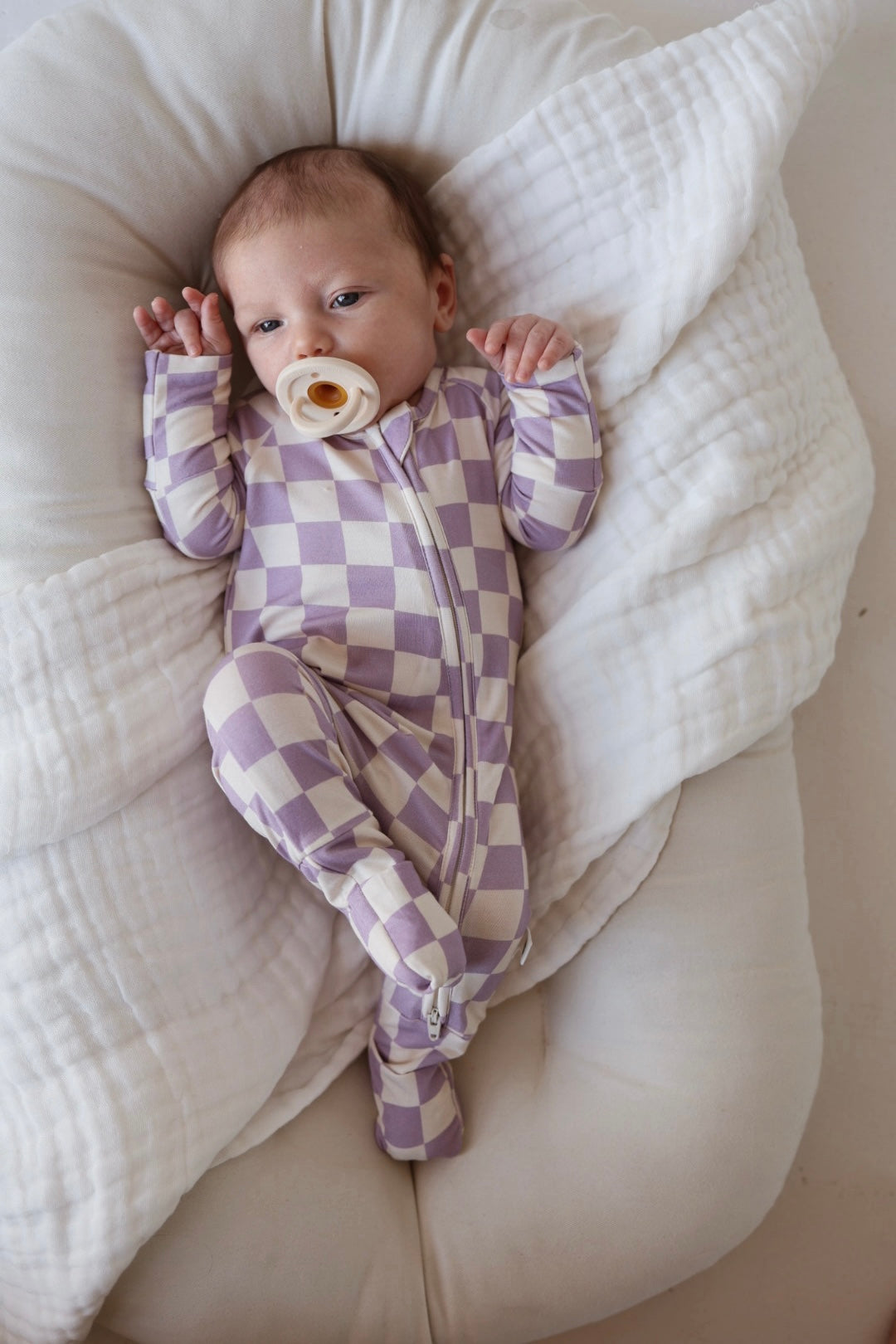 A baby with a pacifier lies on a soft white blanket, wearing the forever french baby Bamboo Zip Pajama in Grape Soda—breathable lavender and white sleepwear—looking up with hands raised near its face.