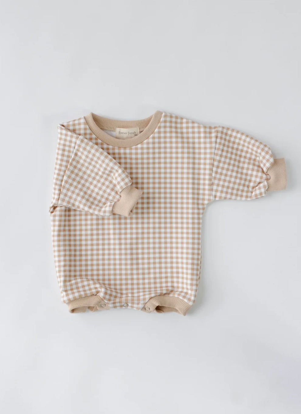 The Goldie Gingham | Sweatshirt Romper by forever french baby, a long-sleeved romper with a beige and white gingham pattern, is displayed on a plain white background. This piece features beige cuffs on the sleeves and leg openings, along with snap buttons at the bottom for fastening.