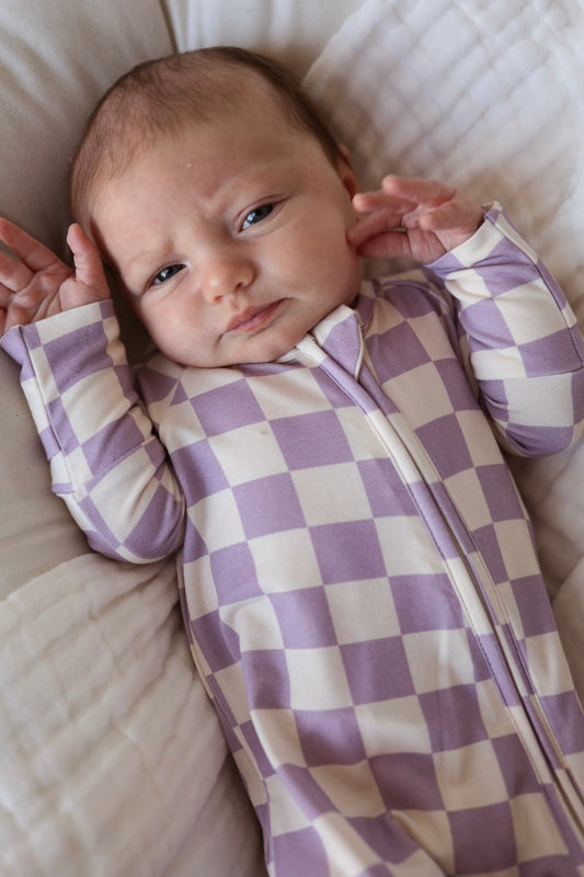 A baby lies on a soft, white blanket wearing forever french baby's Bamboo Zip Pajama in Grape Soda—a breathable zip-up onesie with a light purple and white checkered pattern—looking up with hands near their face.