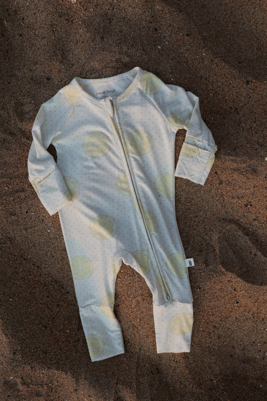The Bamboo Zip Pajamas | Lemon Drop by forever french baby is a light-colored baby onesie featuring yellow lemon slice prints and a front zipper, laid flat on sandy ground—made from breathable fabric for ultimate comfort.