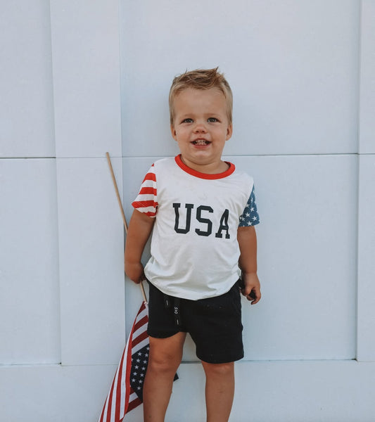 A young child with a messy face and slight smile stands against a white wall, wearing the forever french baby Child Bamboo T-Shirt | Stars & Stripes, black shorts, and holding an American flag.