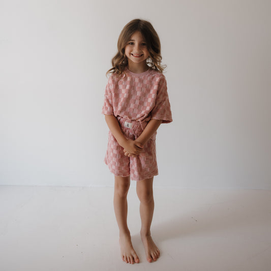 A young girl with wavy brown hair smiles at the camera, barefoot on a white floor, wearing the forever french baby Child Terry Short Set in Sea Pink—ideal for cozy family matching outfits.