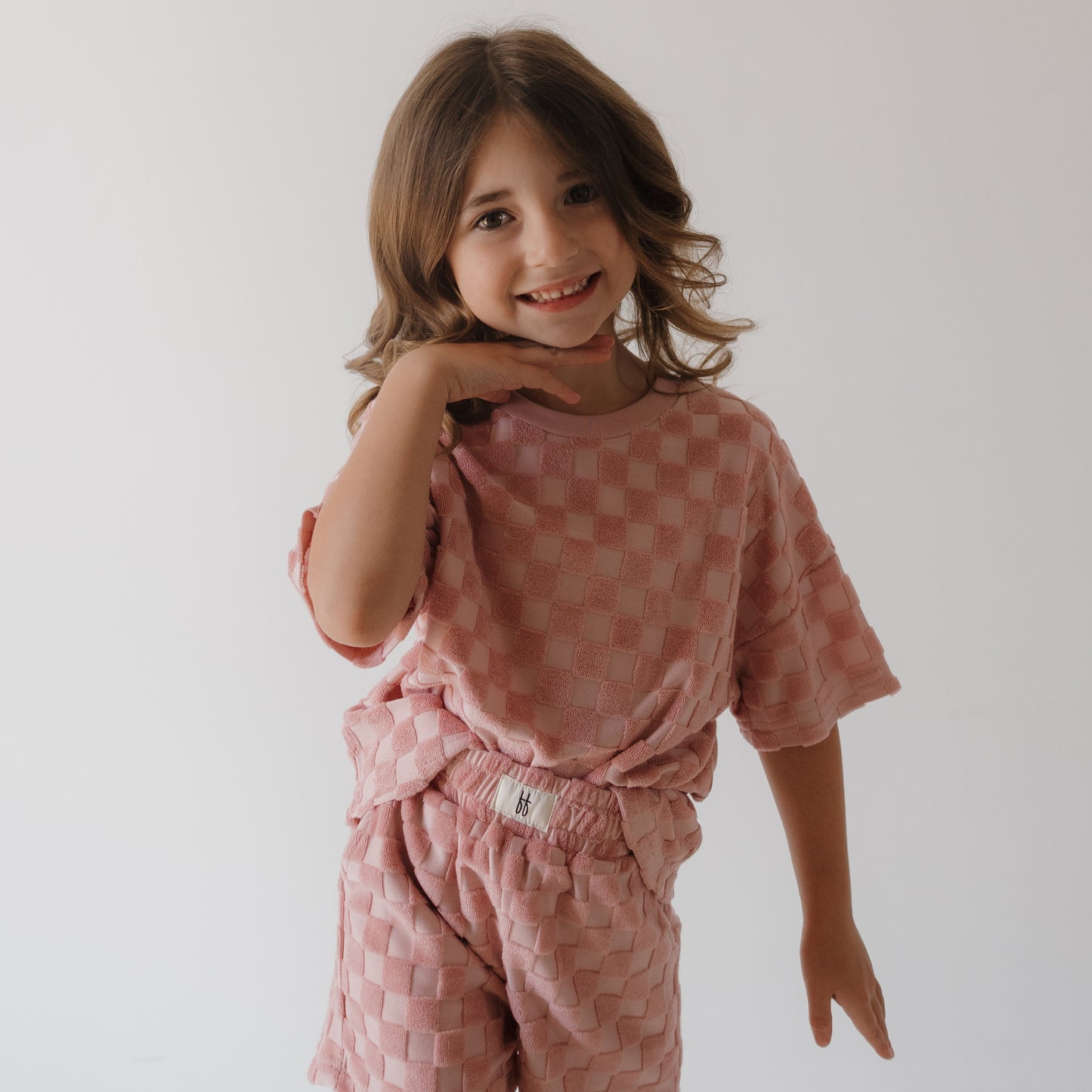 A young child with wavy brown hair smiles and poses with one hand under their chin, wearing the forever french baby Child Terry Short Set in Sea Pink against a plain white background.