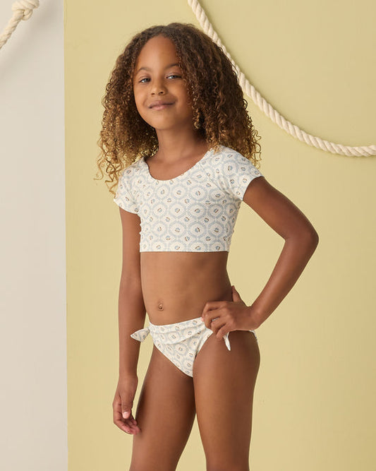 A young girl with curly hair wears the Rylee + Cru Cropped Rashguard Set in Green Fleur, featuring a short-sleeve crop top and matching bikini bottoms with a subtle pattern. She stands in front of a beige wall accented with decorative rope. This swimwear set offers UPF 50+ sun protection, blending style and safety for sunny days.