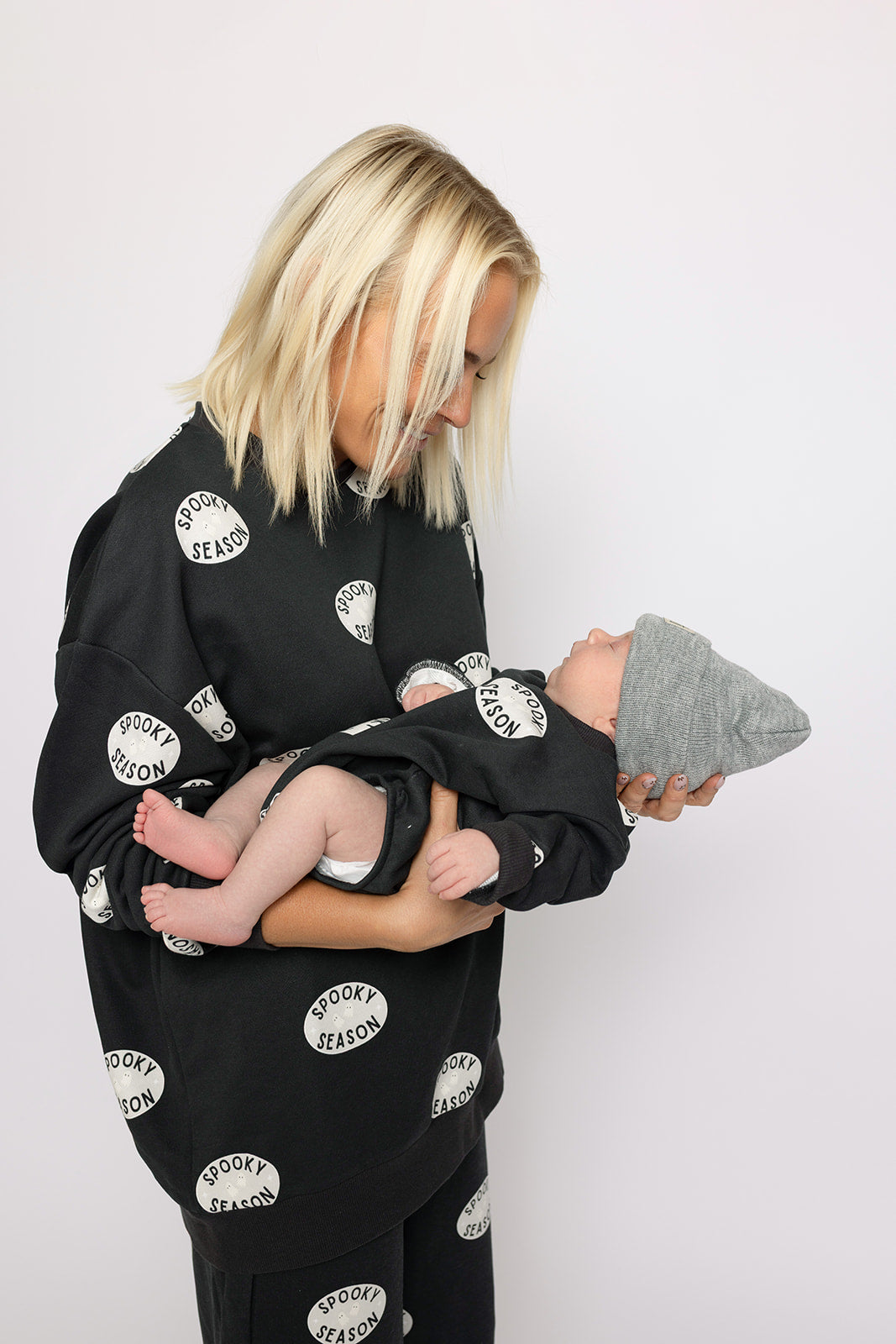A blonde woman smiles holding a baby in a gray hat and Halloween outfit. Both wear matching "Spooky Season" Sweatshirt Romper sets by forever french baby, standing against a plain, light background.