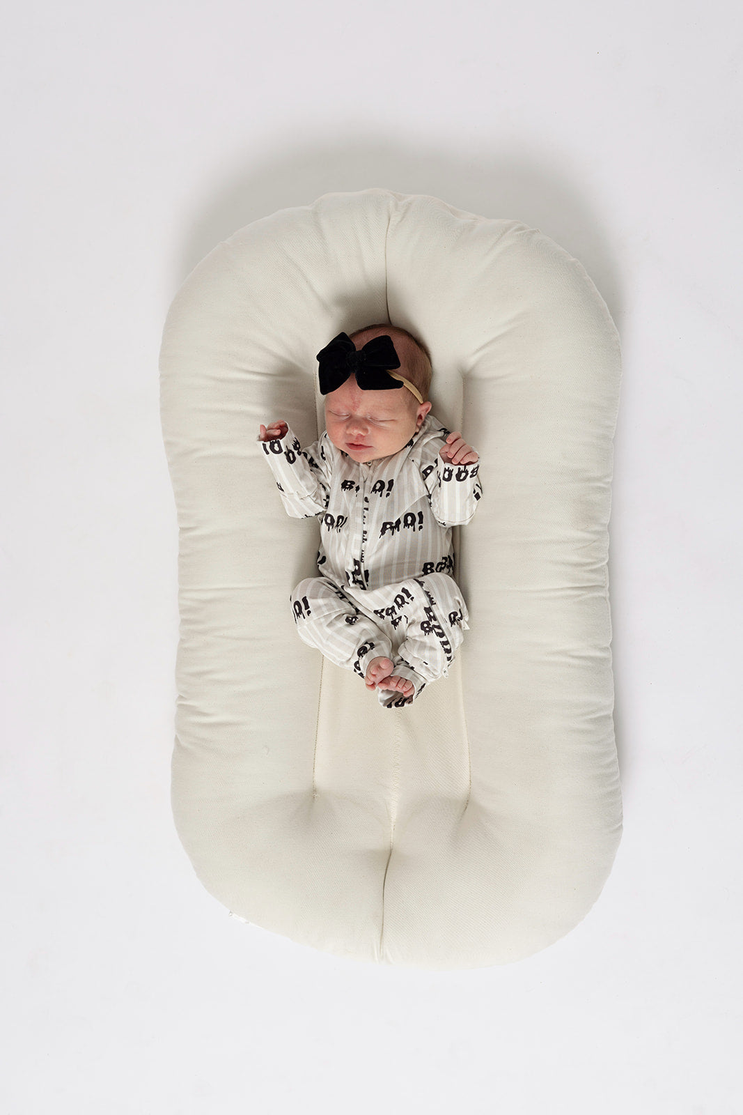 A newborn wearing forever french baby's Bamboo Zip Pajamas | Boo! and a black bow headband lies on a hypoallergenic, oval white cushion against a white background.