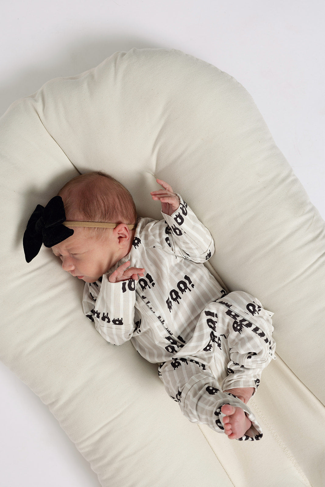 A newborn sleeps peacefully on a soft, white surface, wearing forever french baby Bamboo Zip Pajamas | Boo!—breathable sleepwear featuring black "ROAR" text—and a black bow headband.