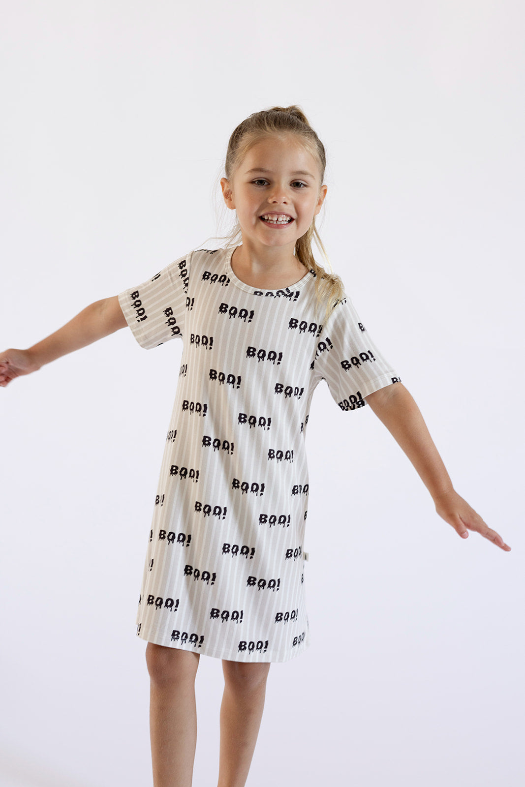 A smiling young girl with outstretched arms wears the forever french baby Bamboo T-Shirt Dress | Boo!, a hypo-allergenic dress featuring "BOO!" and little black ghosts, against a plain white background.
