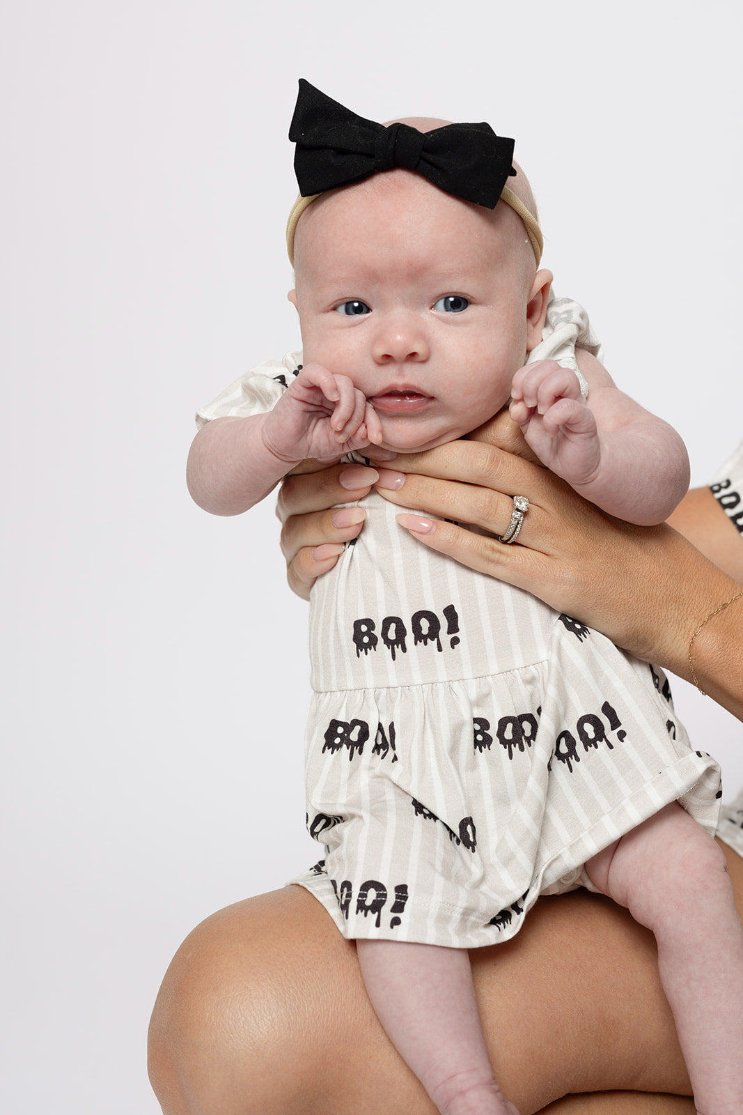 A baby wears the forever french baby Bamboo Ruffle Romper | Boo! in cream with a black "BOO!" print and matching black bow headband, offering hypoallergenic comfort as the baby looks forward with a neutral expression.