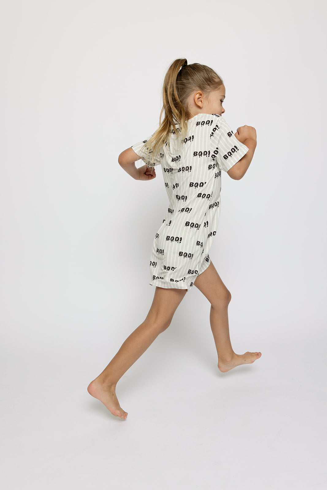 A young girl with blonde hair in a ponytail runs barefoot against a white background, wearing the forever french baby Bamboo T-Shirt Dress | Boo!, a short-sleeved, hypoallergenic dress with a black and white "Boo!" pattern.