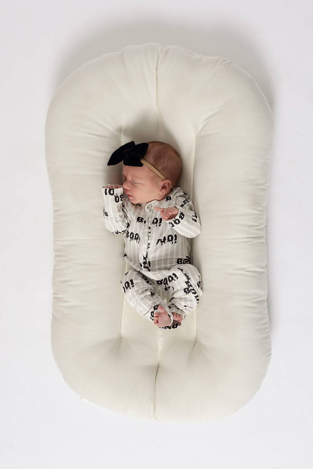 A newborn wearing forever french baby Bamboo Zip Pajamas | Boo!—a white outfit with black text and a black bow headband—lies on a soft, oval-shaped white cushion against a white background.