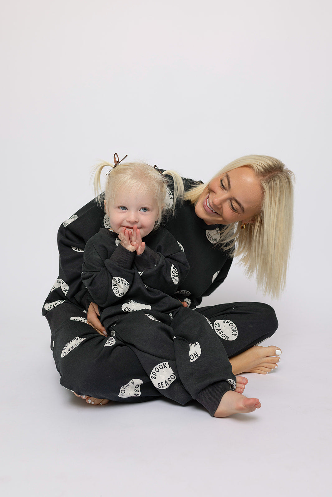 A blonde woman and her daughter smile and pose on the floor in matching forever french baby black "Child Sweat Set | Spooky Season" sweatsuits with white circles, against a plain white background—perfect for spooky season fun!.