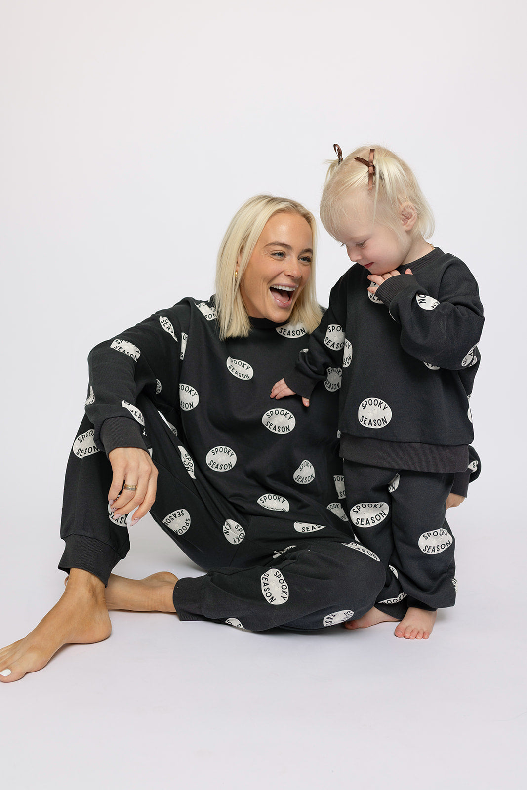 A smiling woman and child wear matching black “Spooky Season” sweat sets from forever french baby, featuring white tooth prints, as they sit together against a plain white background.