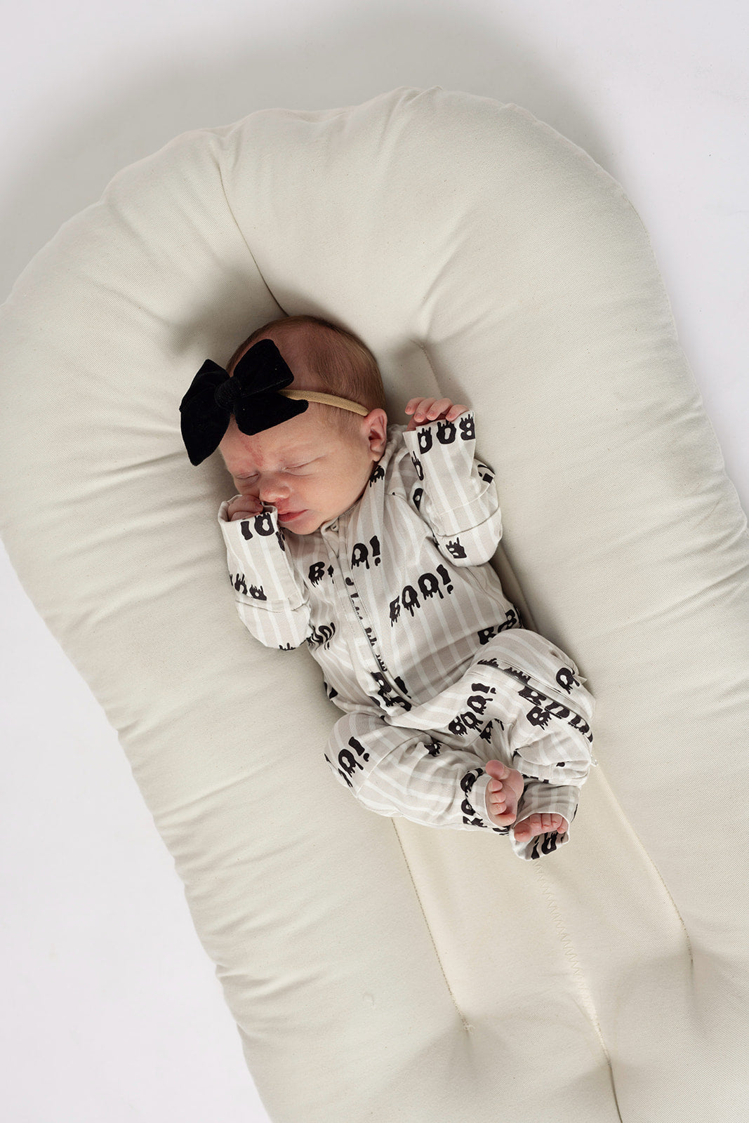 A newborn sleeps peacefully on a soft white pad, dressed in forever french baby's Bamboo Zip Pajamas | Boo!—a hypoallergenic, white onesie with black patterns and a large black bow headband.