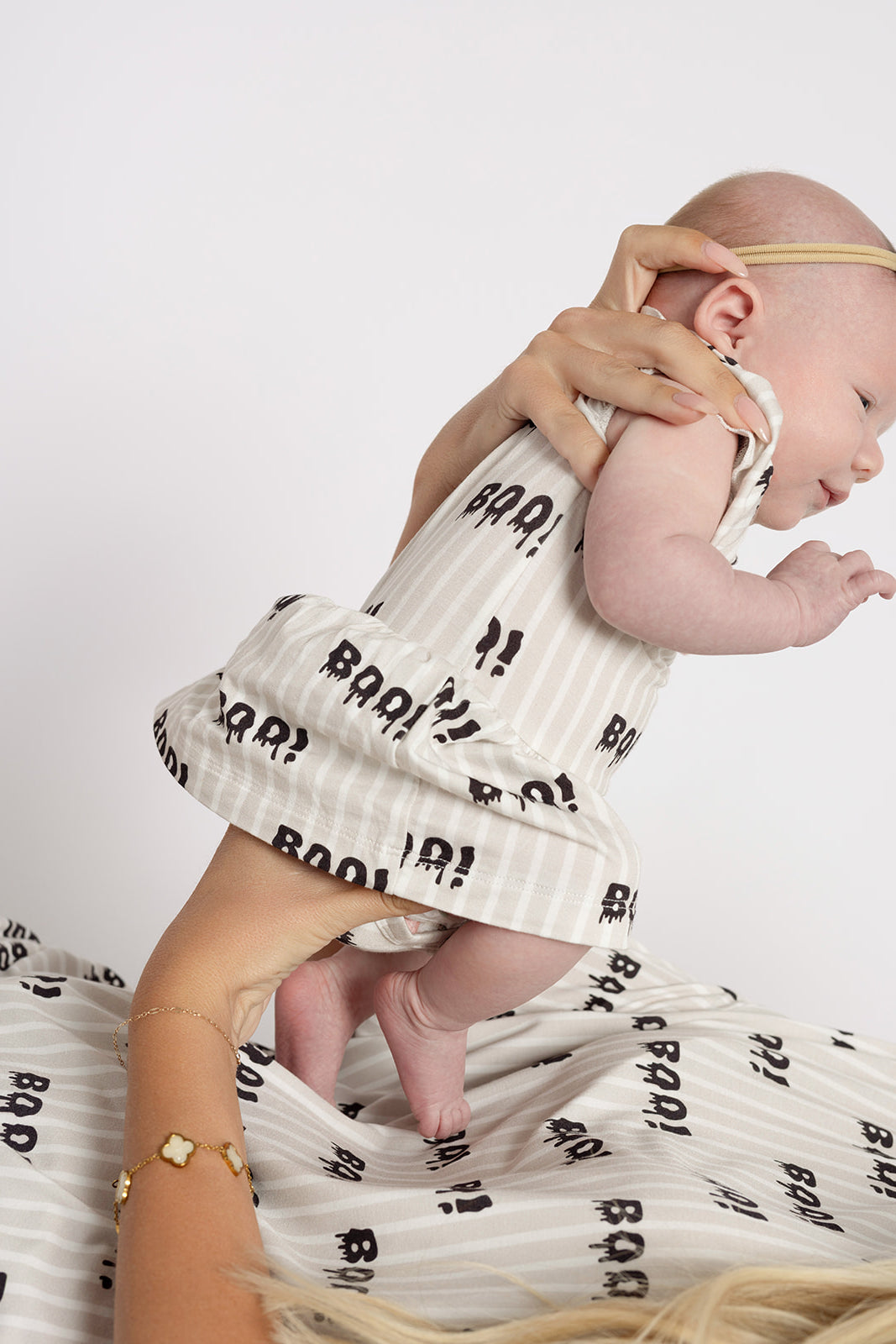 A baby in a cream Bamboo Ruffle Romper | Boo! from forever french baby, featuring black “Boo!” text, is held by an adult in matching print with gold bracelets. The soft, hypoallergenic romper pops against the white background.