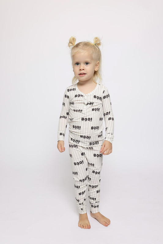 A young child with blonde hair in two buns stands barefoot on a white background, wearing forever french baby's Bamboo Two Piece Pajamas | Boo!—breathable children’s sleepwear featuring black "BOO!" prints for comfort and style.