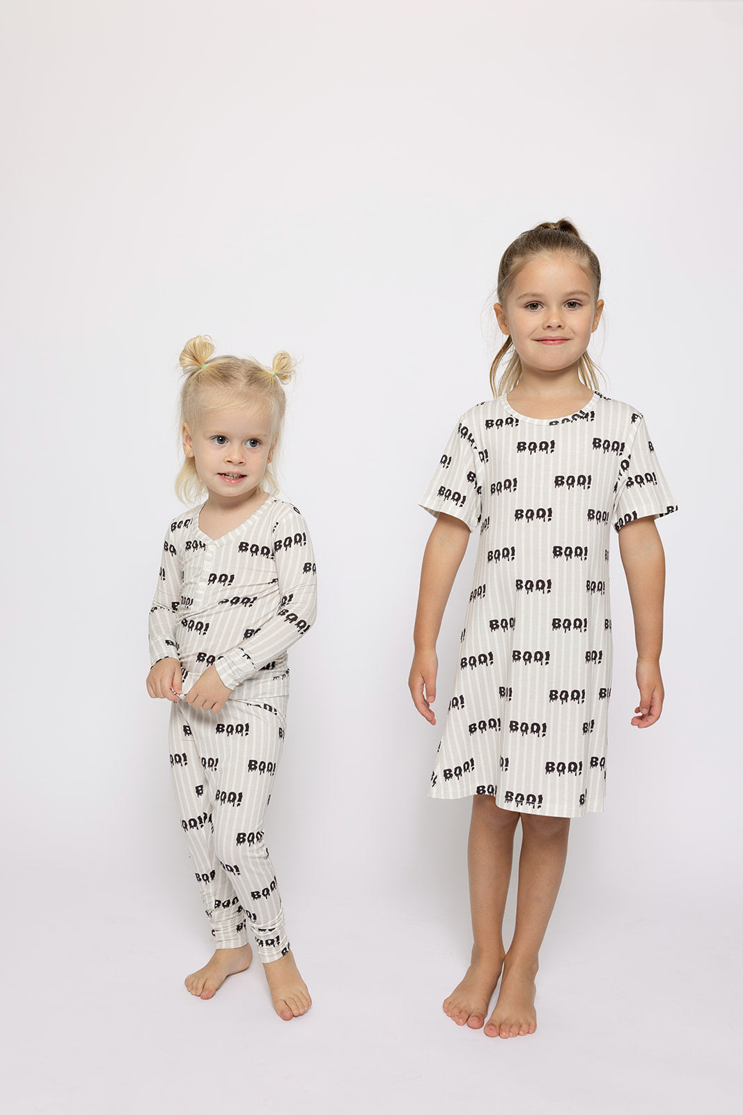 Two young girls stand barefoot against a white background in matching, breathable forever french baby Bamboo Two Piece Pajamas | Boo!—one wears the pants set, the other a short-sleeved nightgown—both with a fun black "BOO!" print.