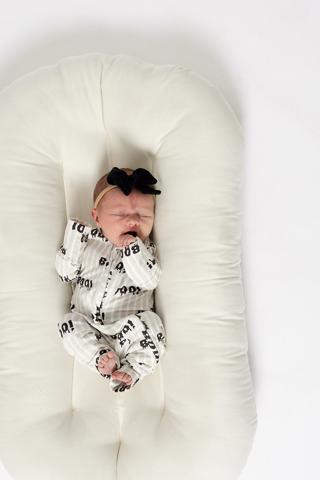 A newborn sleeps peacefully in forever french baby Bamboo Zip Pajamas | Boo!—a striped outfit with a black bow headband—on a soft, white, oval cushion against a white background.