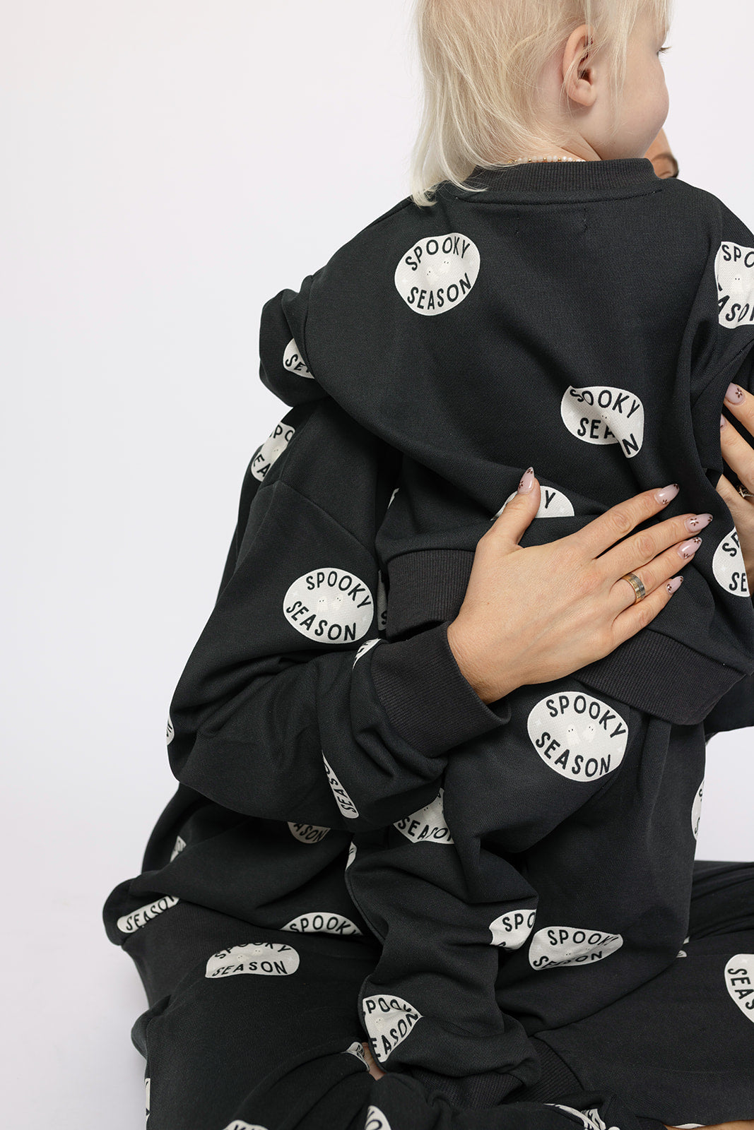 Two people in matching black forever french baby "Spooky Season" Child Sweat Sets sit close, one hugging the other and displaying their hands and rings. Their faces are mostly hidden, ideal for a cozy matching look this spooky season.