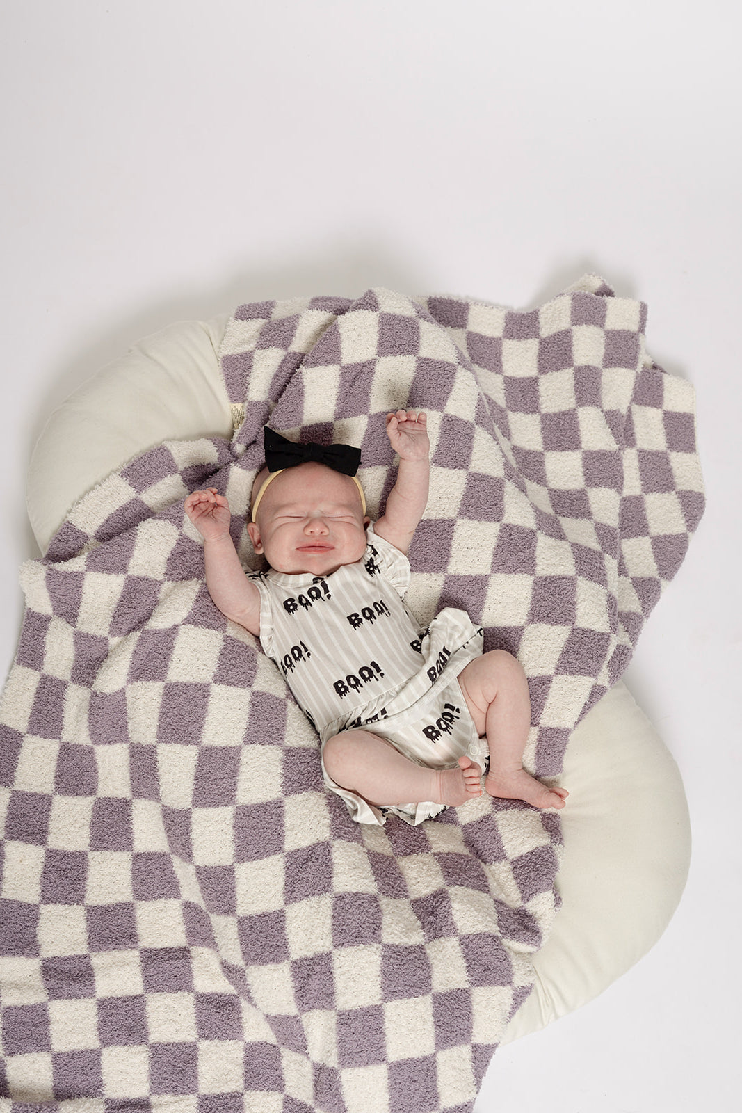 A baby in a forever french baby Bamboo Ruffle Romper | Boo! featuring "ROAR" text and a black bow headband lies on a purple and white checkered blanket—an adorable, hypoallergenic outfit choice.