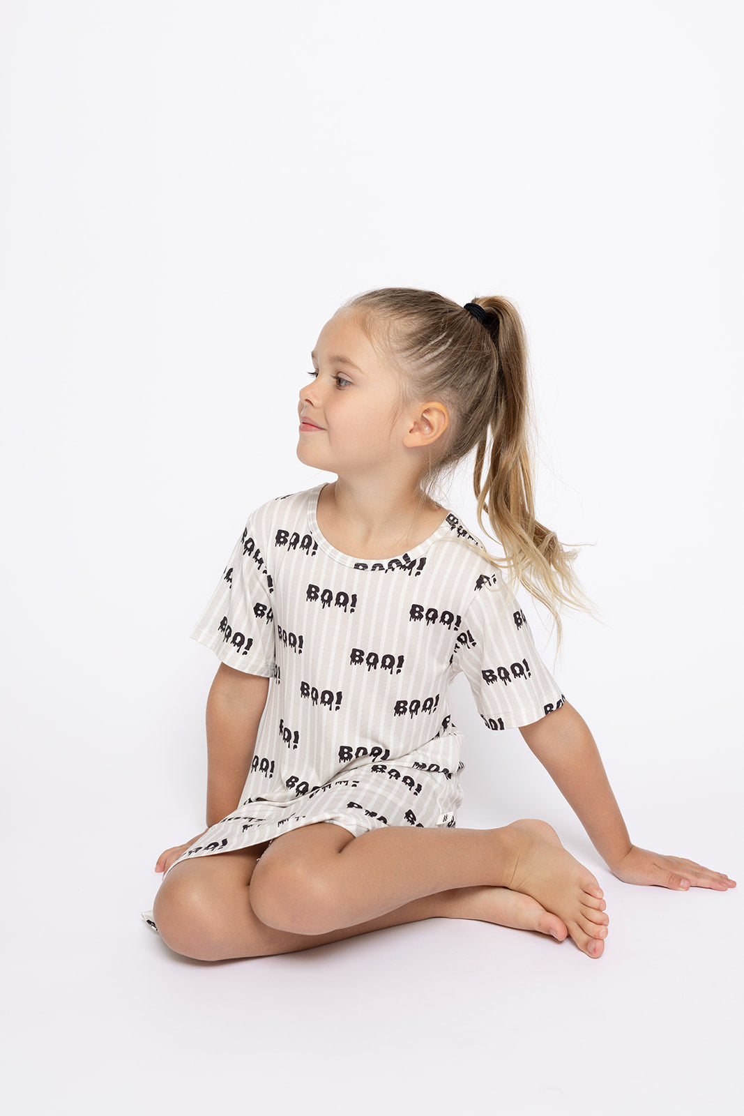 A young girl with long blonde hair in a ponytail sits cross-legged, wearing the forever french baby Bamboo T-Shirt Dress | Boo! featuring a black "BOO!" print, against a plain white background.