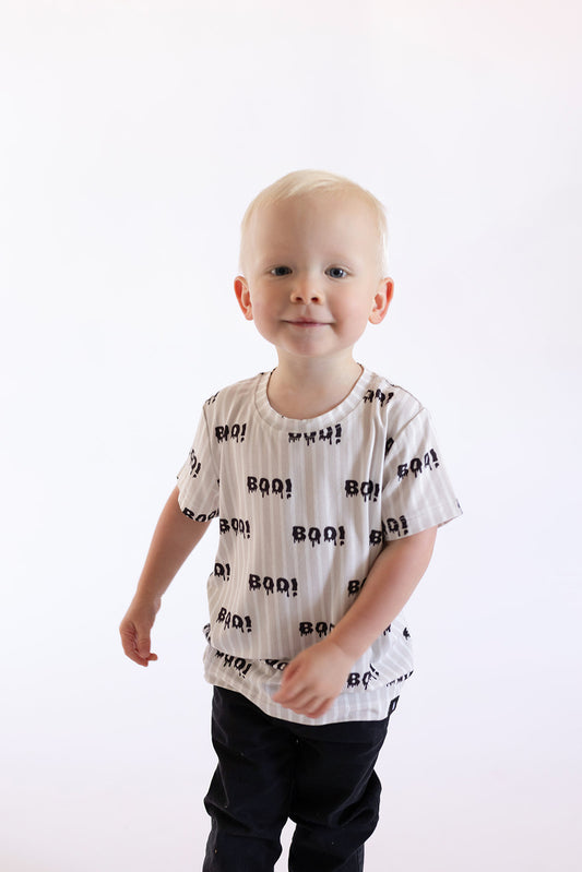A young child with short blond hair smiles in a forever french baby Child Bamboo T-Shirt | Boo!—a breathable kids' tee with a black "BOO!" pattern—paired with black pants, standing against a plain white background.