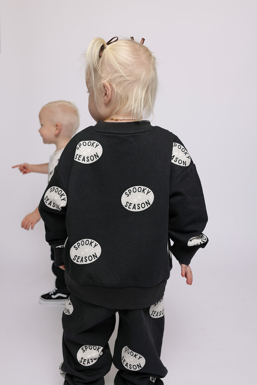 Two young toddlers with blonde hair stand together. One faces away, wearing the forever french baby Child Sweat Set | Spooky Season in black. The other points outward, dressed in lighter clothes against a plain white background.