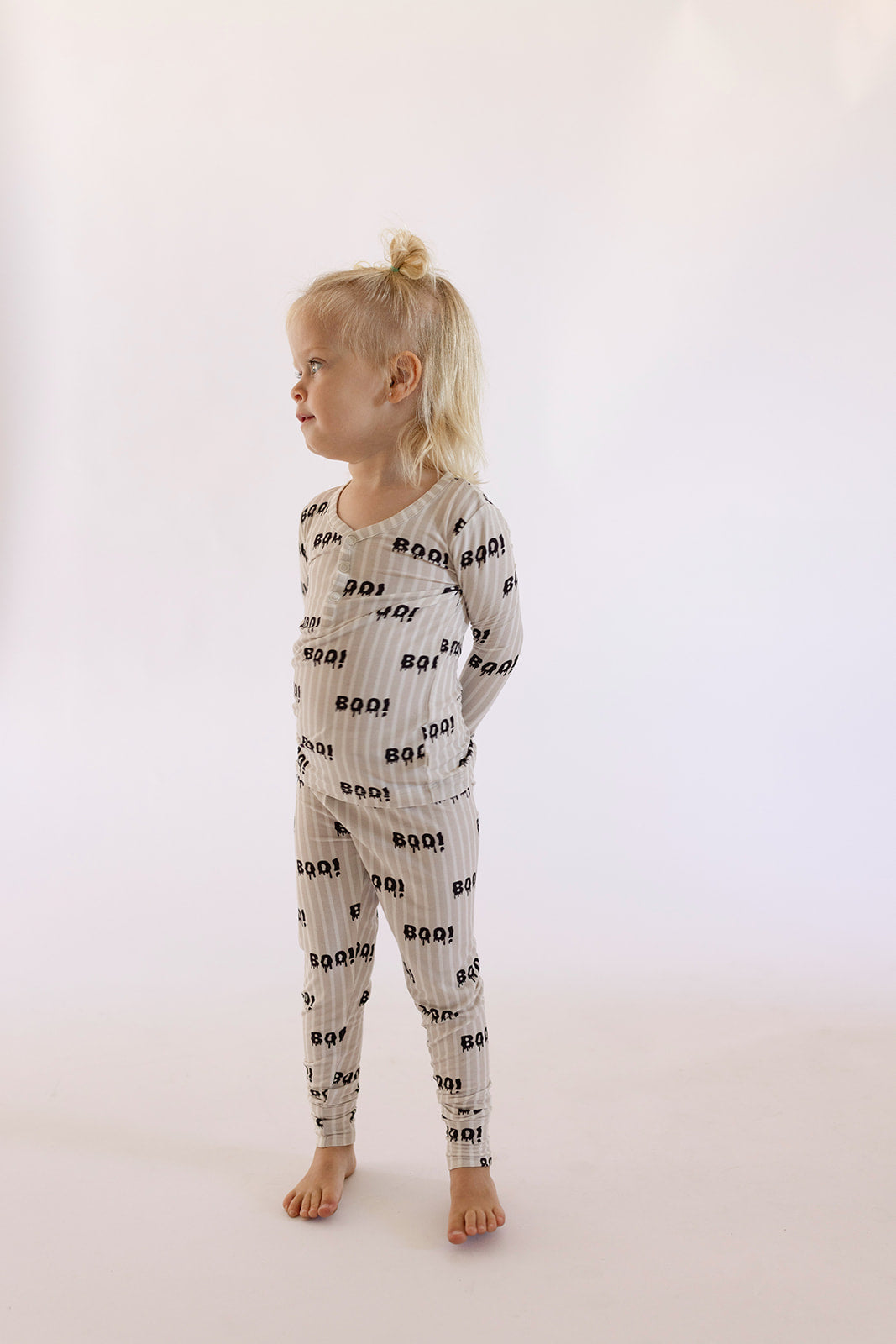 A young child with light blonde hair in a topknot stands barefoot against a plain light background, wearing forever french baby's Bamboo Two Piece Pajamas | Boo! featuring matching long sleeves and pants with a repeating "BOO!" print.
