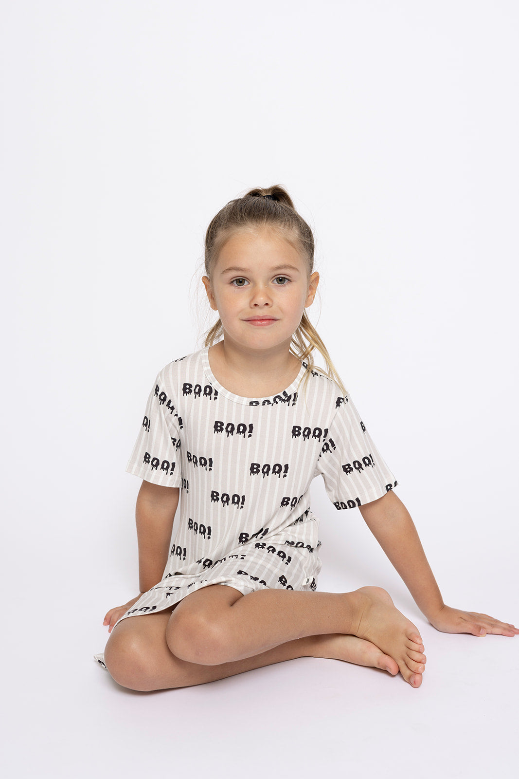 A young girl with light brown hair in a ponytail sits barefoot against a white background, wearing the forever french baby Bamboo T-Shirt Dress | Boo!, a short-sleeved, hypo-allergenic dress featuring a playful black "boo!" pattern.
