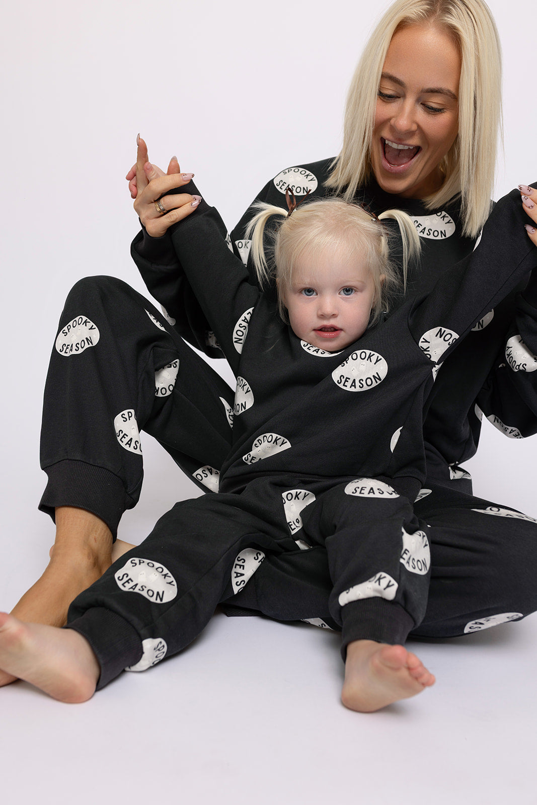 A smiling woman and her daughter pose in matching “Spooky Season” black pajamas. The child, sporting blonde pigtails, wears the forever french baby Child Sweat Set | Spooky Season against a plain white background.
