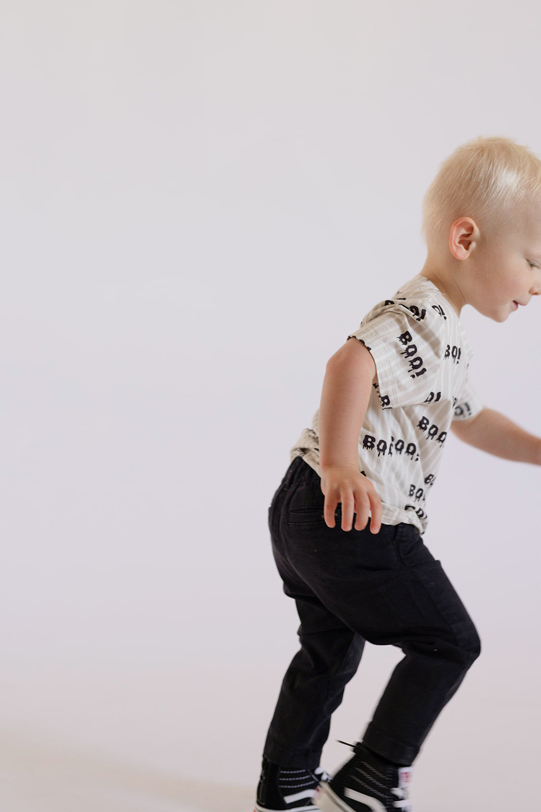 A young child with short blond hair, wearing a forever french baby Child Bamboo T-Shirt | Boo! and dark pants, is walking or playing against a plain white background.