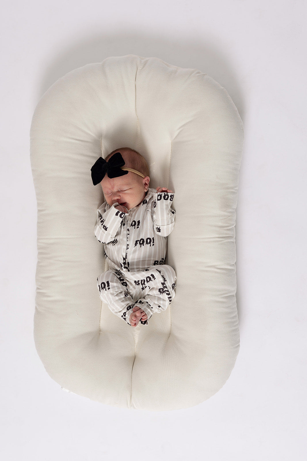 A newborn in forever french baby Bamboo Zip Pajamas | Boo!—a white outfit with black patterns and a large black bow headband—rests peacefully on a soft, oval-shaped white cushion, hands near their face.