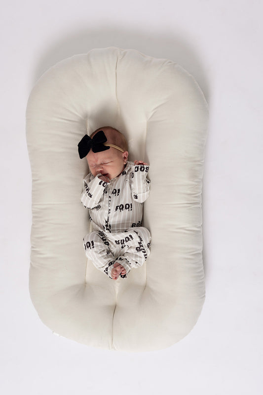 A newborn in forever french baby Bamboo Zip Pajamas | Boo!—a white outfit with black patterns and a large black bow headband—rests peacefully on a soft, oval-shaped white cushion, hands near their face.
