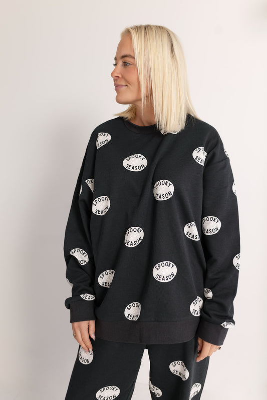 A woman with straight blonde hair wears the forever french baby Women's Sweat Set | Spooky Season—black with white "Spooky Season" ovals. She stands against a light background, smiling and looking left.