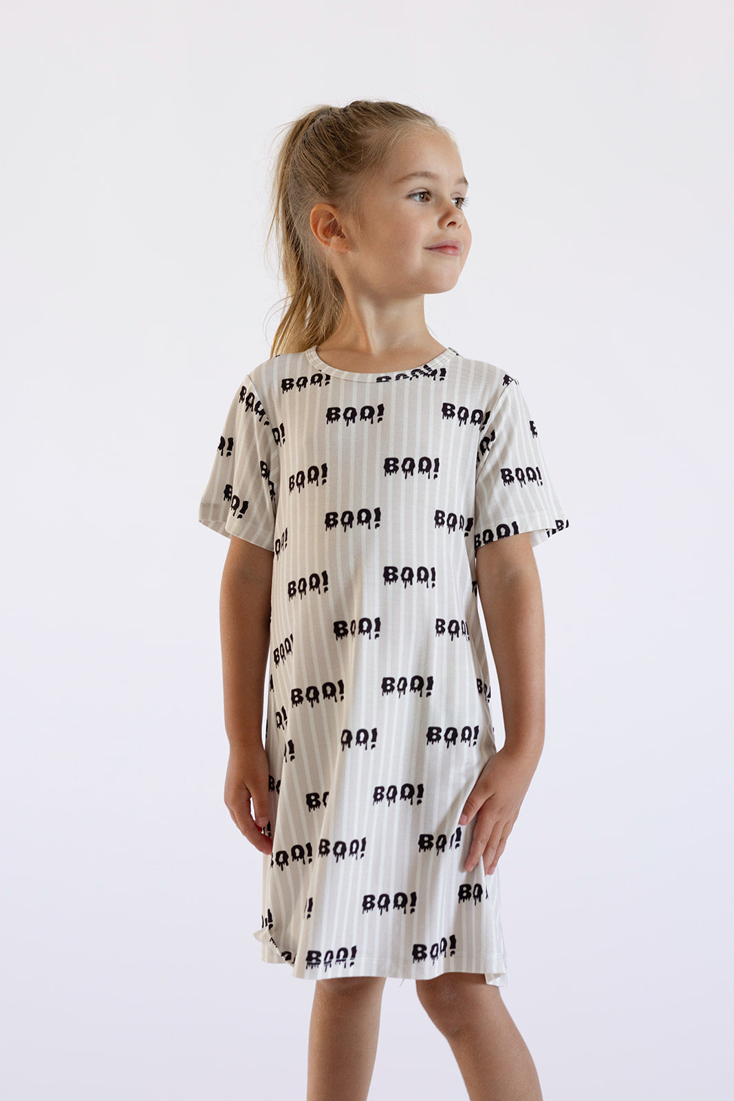 A smiling young girl with blonde hair in a ponytail wears the forever french baby Bamboo T-Shirt Dress | Boo!, featuring “BOO!” text and ghost faces, while standing against a plain light background.