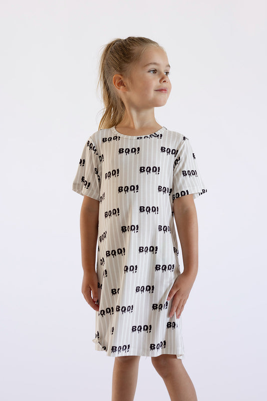 A smiling young girl with blonde hair in a ponytail wears the forever french baby Bamboo T-Shirt Dress | Boo!, featuring “BOO!” text and ghost faces, while standing against a plain light background.