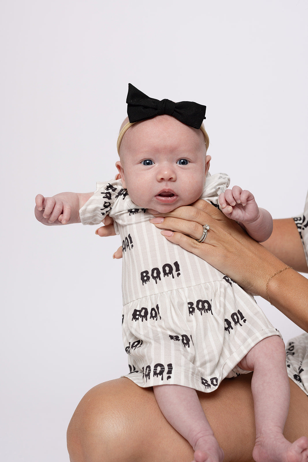 A baby in a forever french baby Bamboo Ruffle Romper | Boo!—a cream hypoallergenic outfit—paired with a black bow headband, is held securely by an adult against a plain light background.