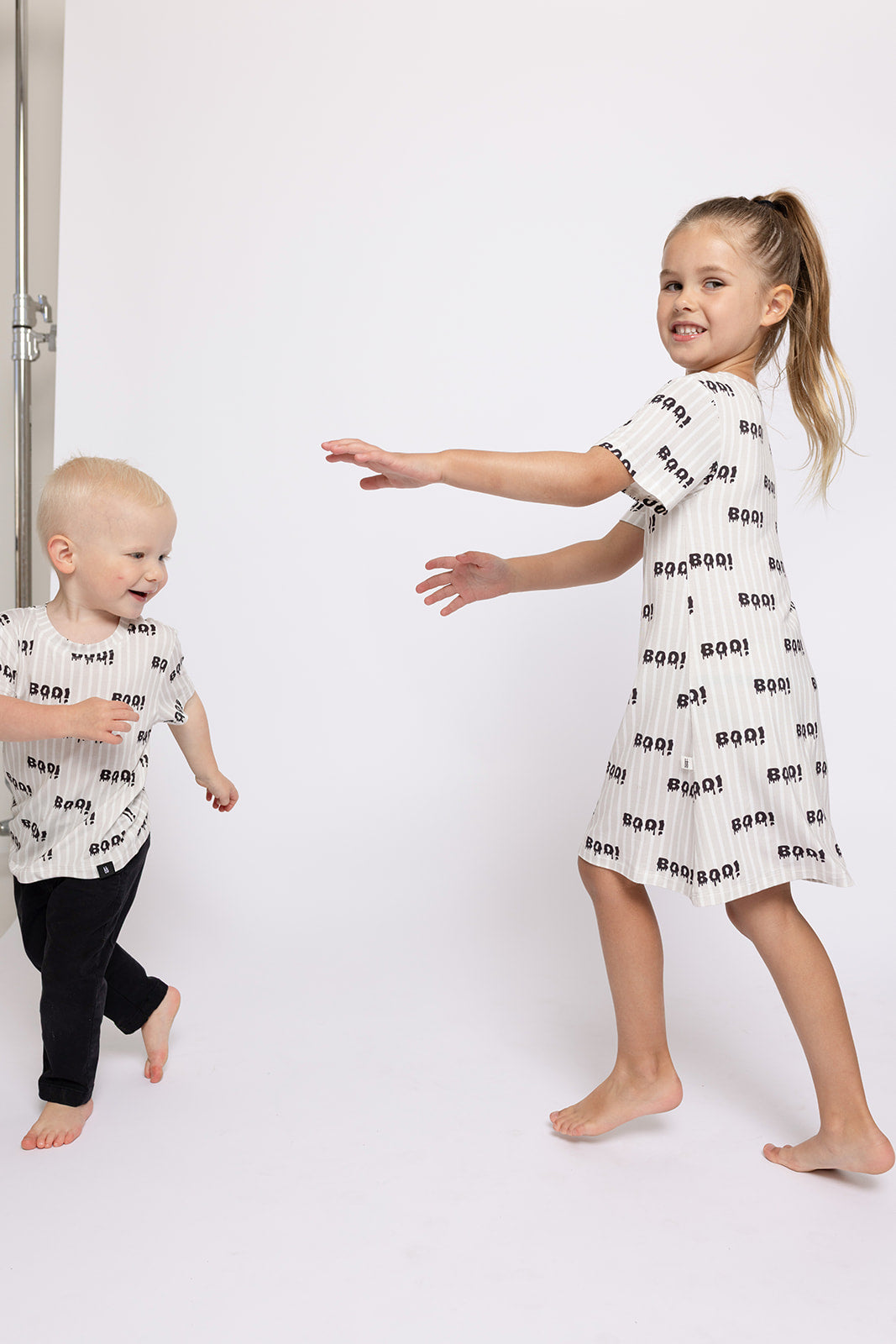 Two kids play indoors against a white background. The girl wears the forever french baby Bamboo T-Shirt Dress | Boo! and both are in matching hypo-allergenic "BOO!" outfits, keeping them comfy as they laugh together.