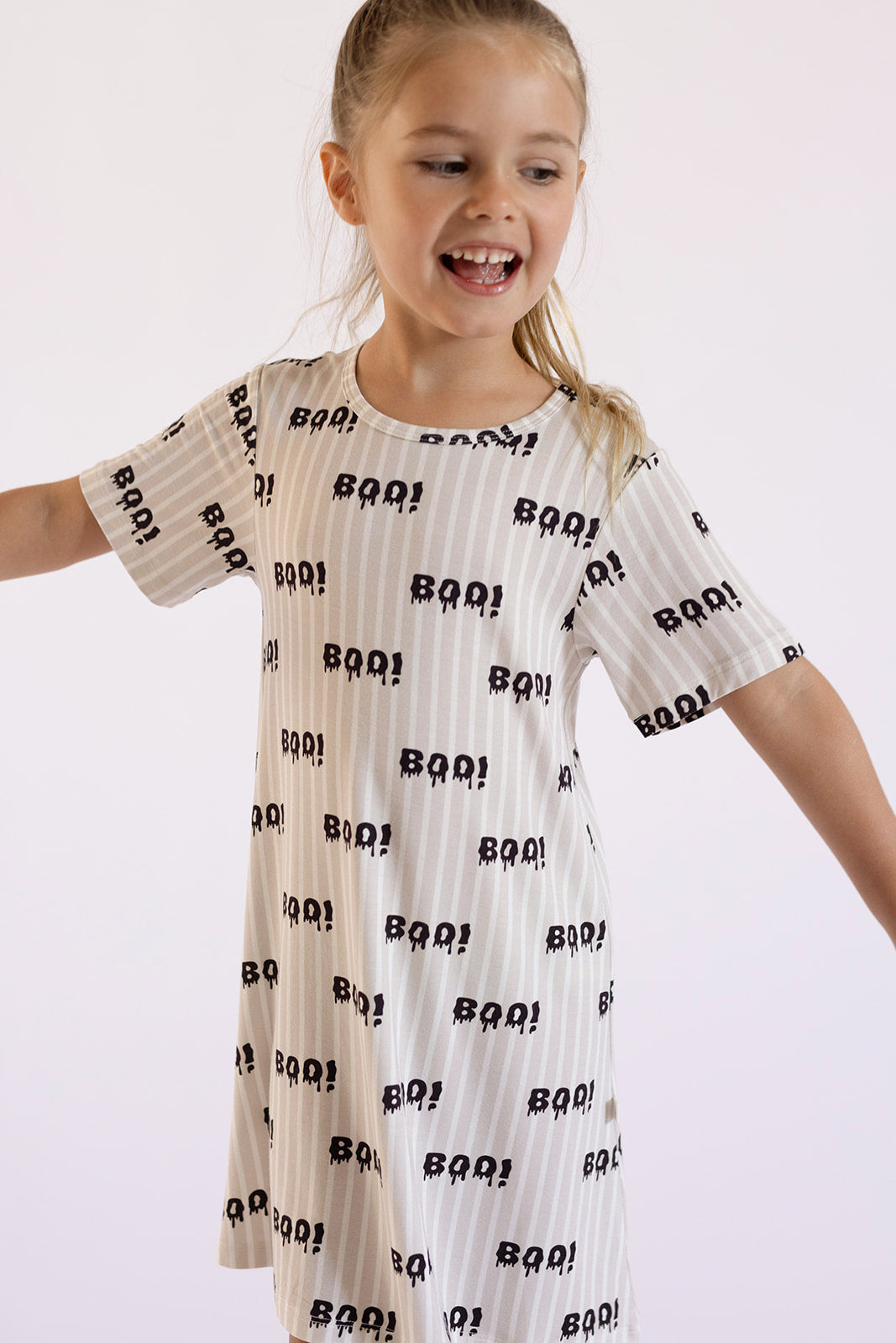 A young girl smiles with arms outstretched in the forever french baby Bamboo T-Shirt Dress | Boo!—a white, hypo-allergenic dress featuring subtle stripes, playful ghost prints, and bold black "BOO!" text, set against a light background.